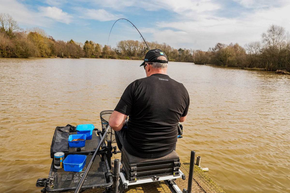 Preston Lightweight Black Raglan T-shirt – achteraanzicht van visser in zwart T-shirt op een zitkist aan het water