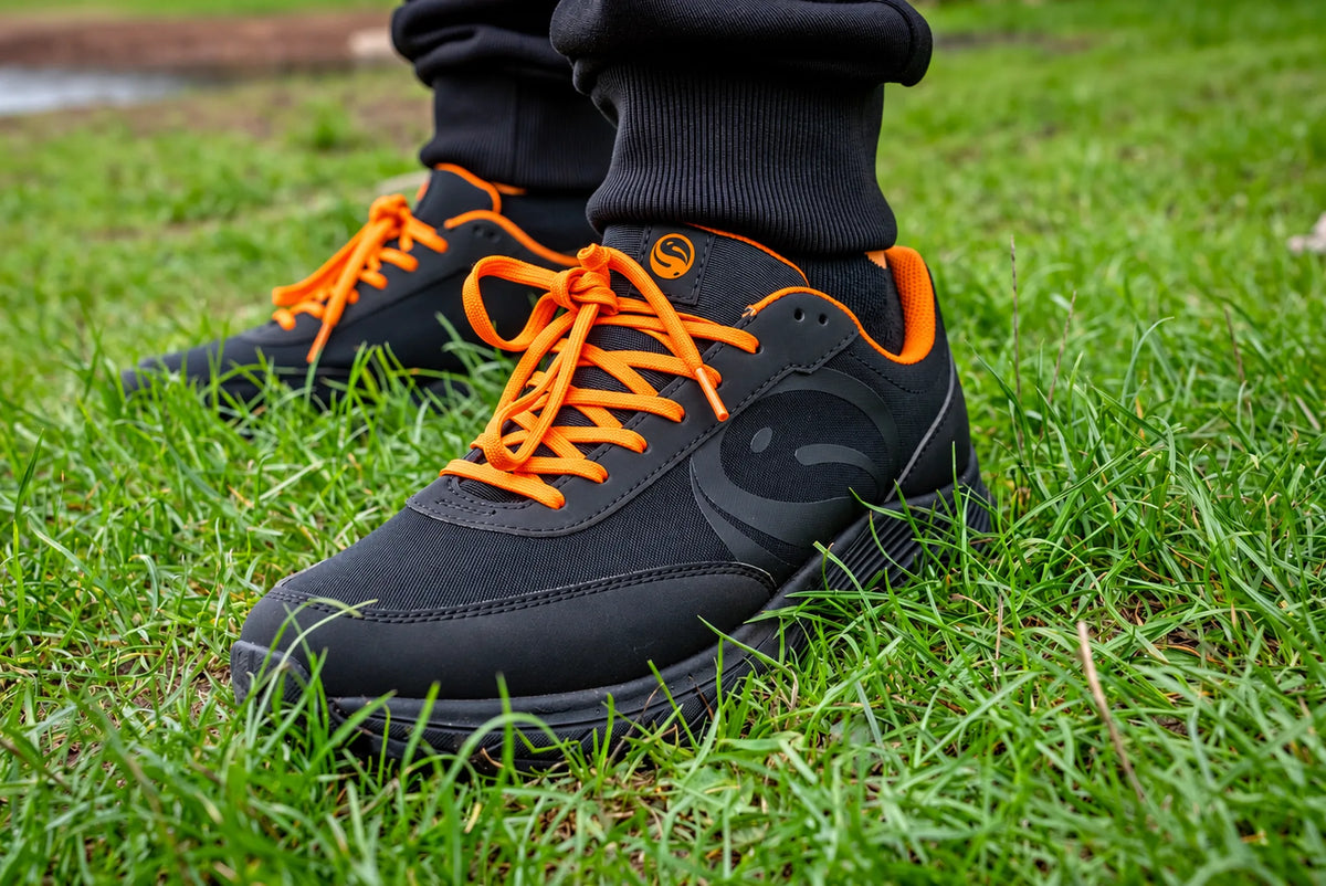 Guru Waterproof Trainer Black – Schuhpaar mit orangefarbenen Schnürsenkeln im Gras