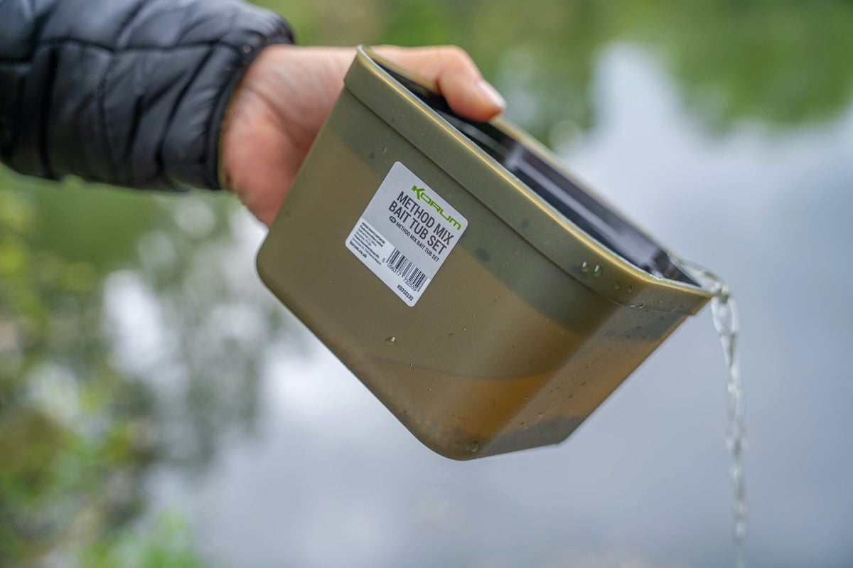 Schuin gehouden Korum Method Mix aasbak met zichtbaar origineel etiket terwijl water langs de rand wegloopt