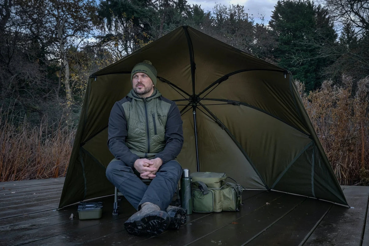 Korum Progress Graphite Brolly Shelter 50 inch – visser zit onder de shelter aan de waterkant