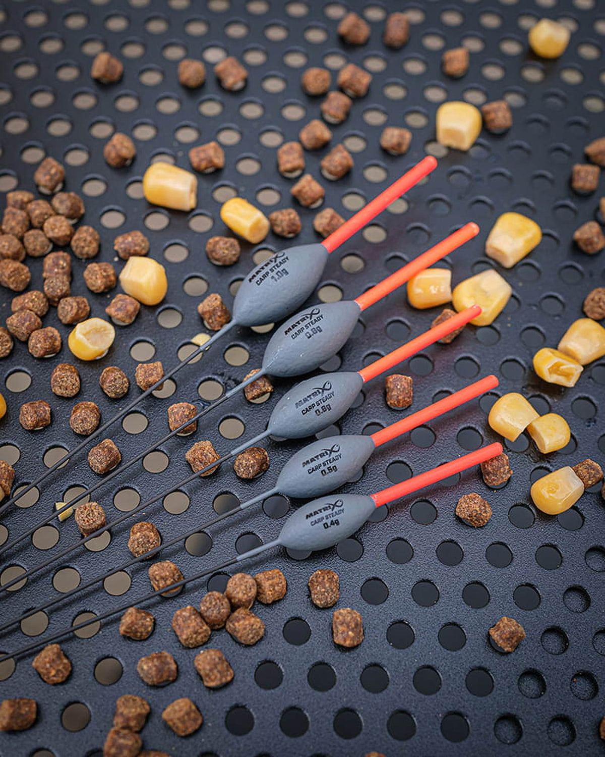 Matrix Carp Steady Pole Floats – vijf dobbers met leesbare gewichten tussen pellets en maïs op een geperforeerde aastafel