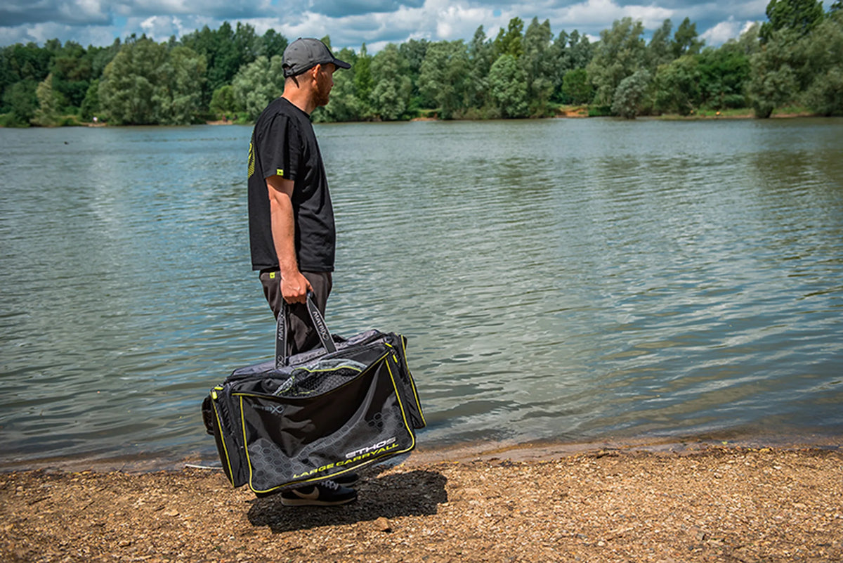 Matrix Ethos Large Carryall – visser draagt de tas met netten in het voorvak aan de handgrepen langs het water