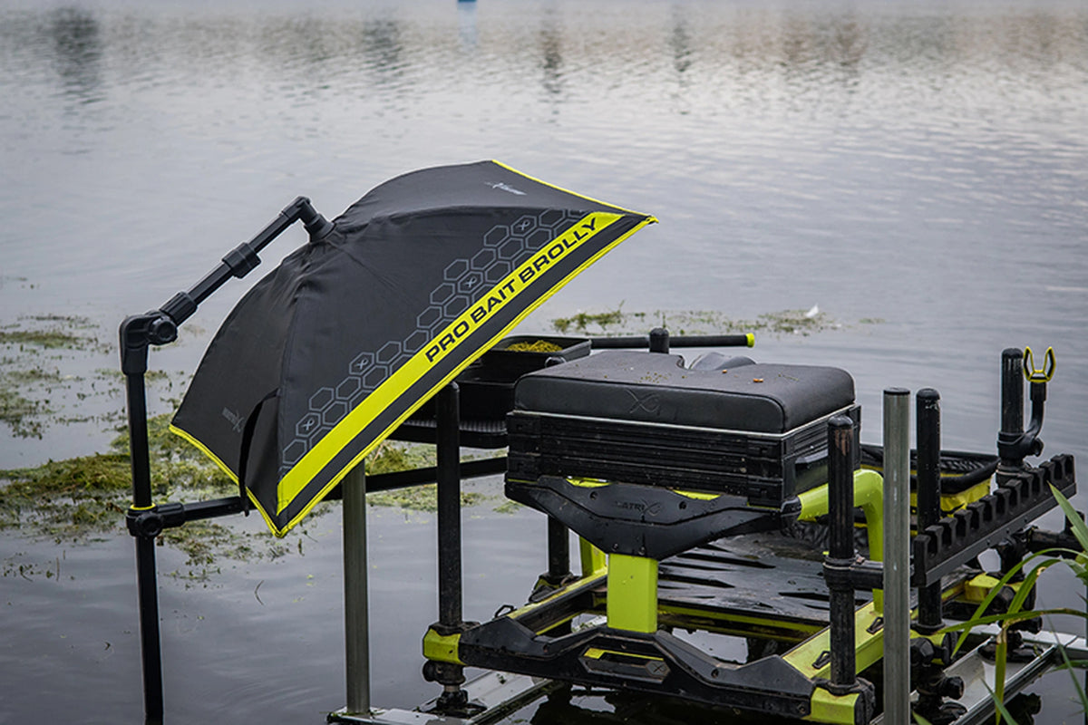 Matrix Pro Bait Brolly aasparaplu – schuin opgesteld naast een zwarte zitkist met gele onderdelen aan de waterkant