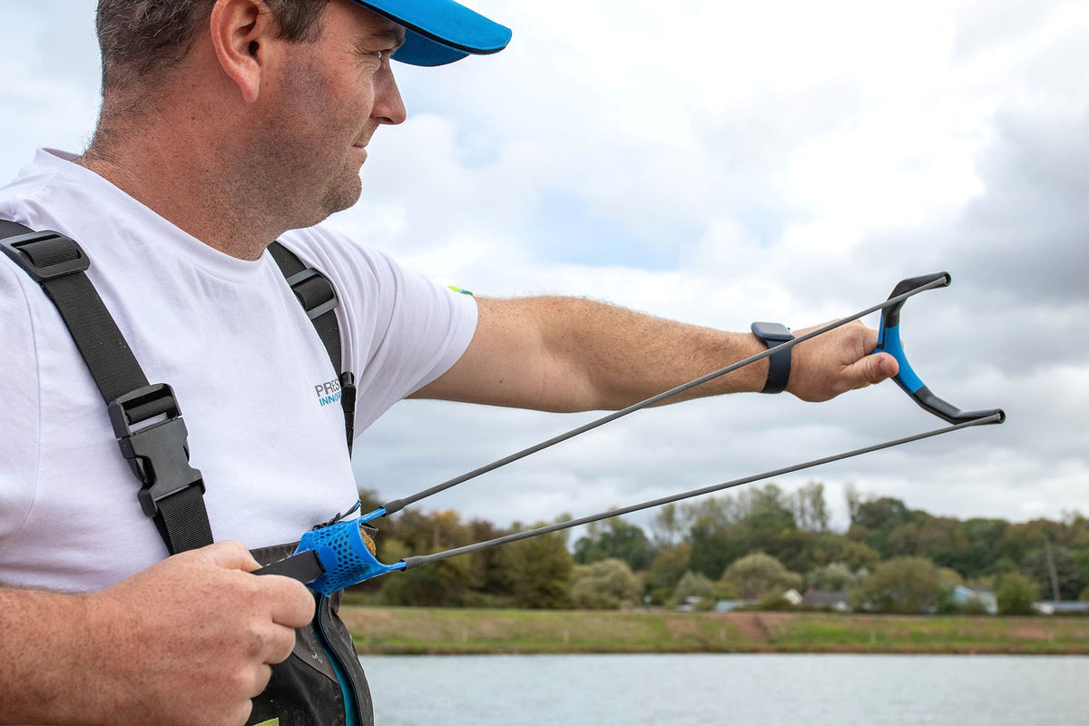 Zijaanzicht van een visser die de Preston Groundbait Catapult met gespannen elastieken op het water richt
