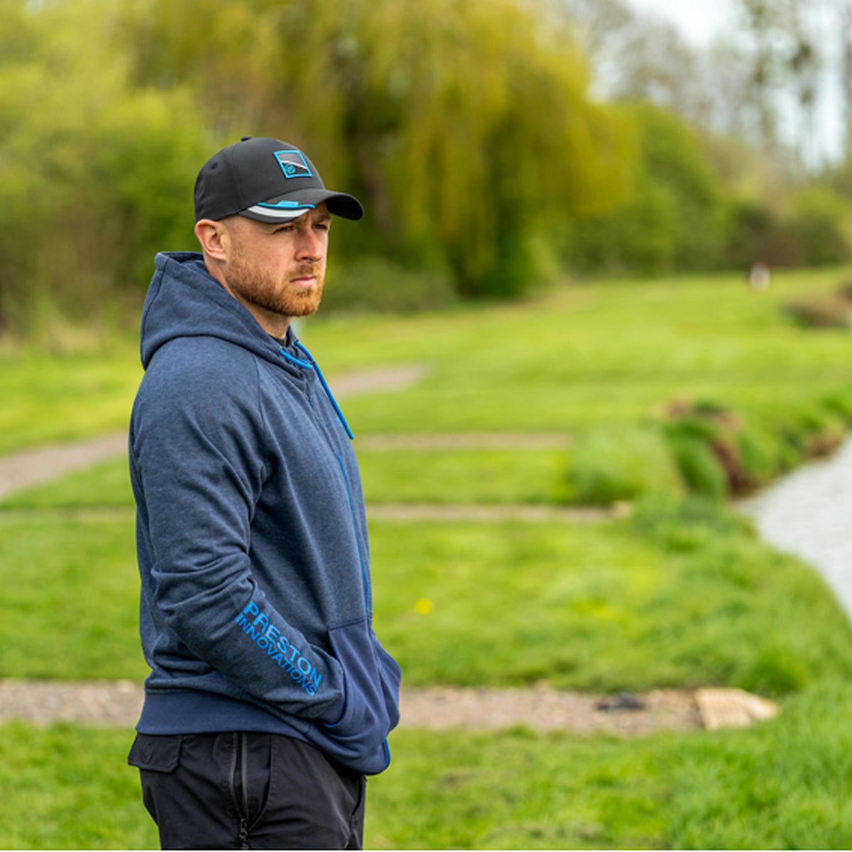 Preston Navy Tide Zip Hoodie – gedragen in zijaanzicht aan de waterkant met capuchon op de rug en handen in de zakken