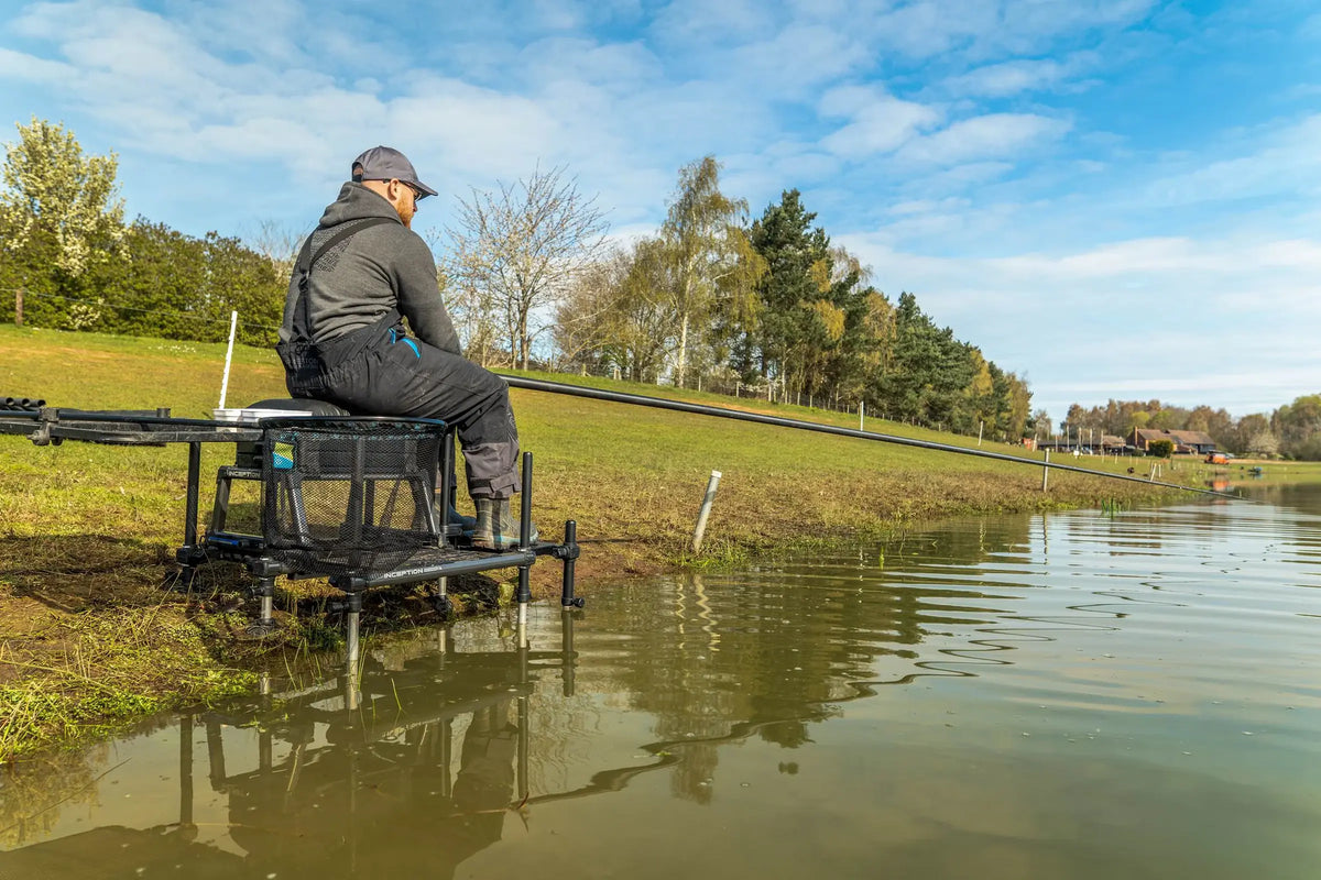 Preston Offbox Side Foot Rest in gebruik naast een zitkist, met een visser aan de waterkant en zijn voeten op de zijvoetensteun