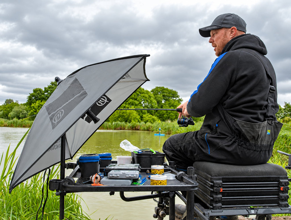 Visser op een zitkist met aastafel op de Preston steunarm en een grijze paraplu aan de waterkant
