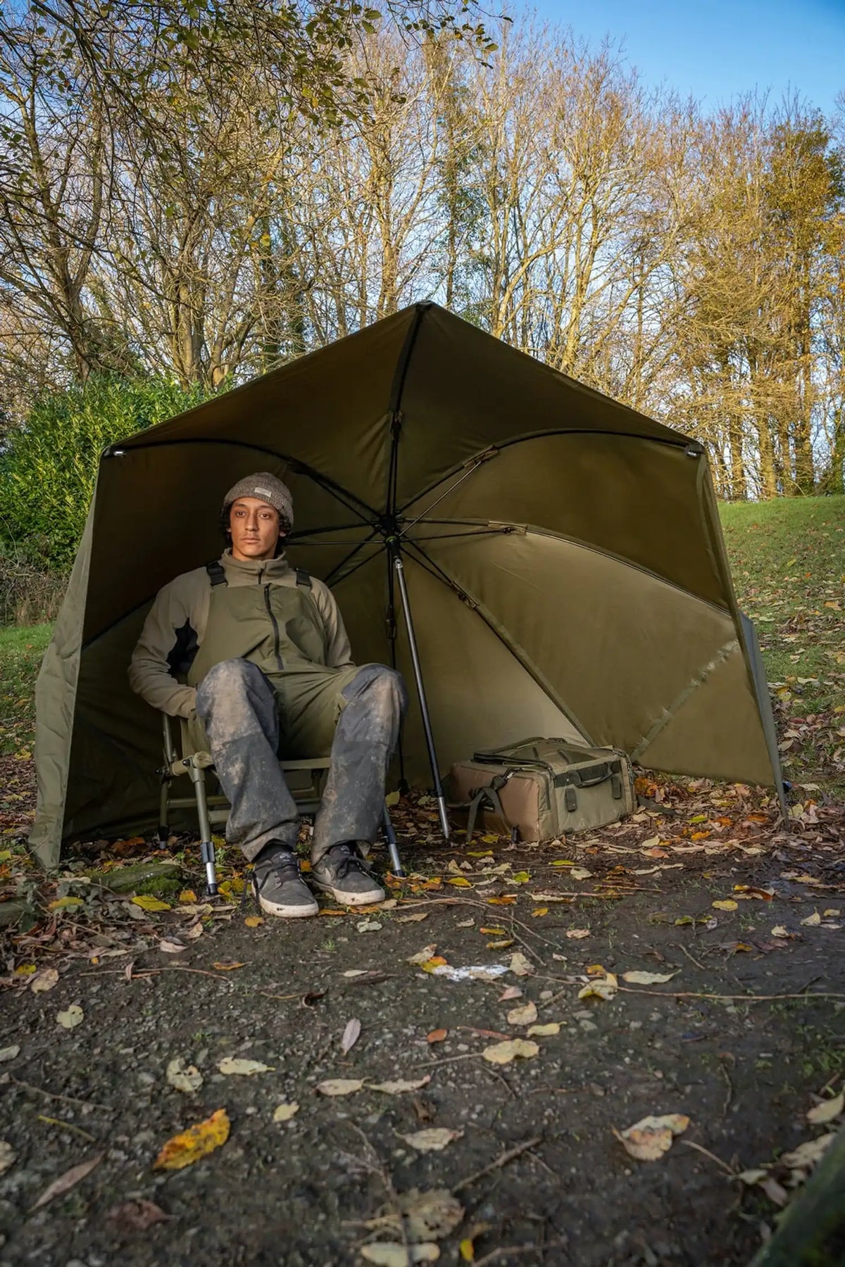 Korum Progress Graphite Brolly Shelter 50 inch – visser zit met tas onder de groene shelter