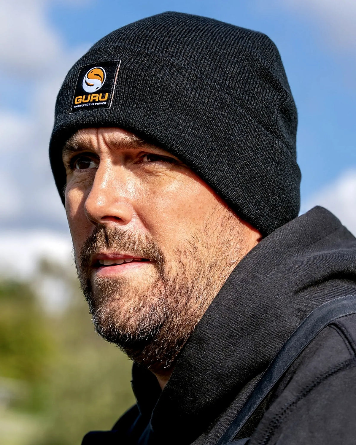 Guru Black Patch Beanie – gedragen in de buitenlucht met blauwe lucht achter de zwarte gebreide muts