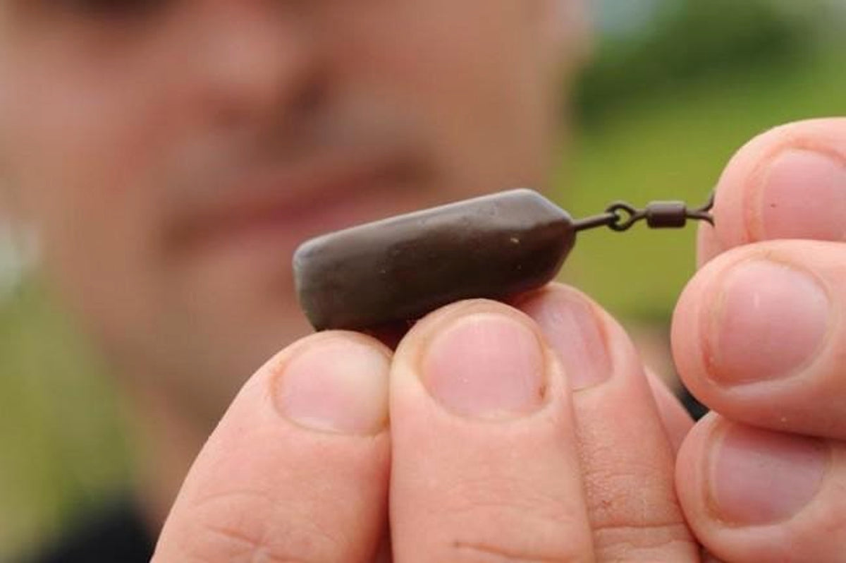 Zijaanzicht van een bruine Guru Flat Pear Bomb tussen vingers, met wartel en klein rubber bij de lijn