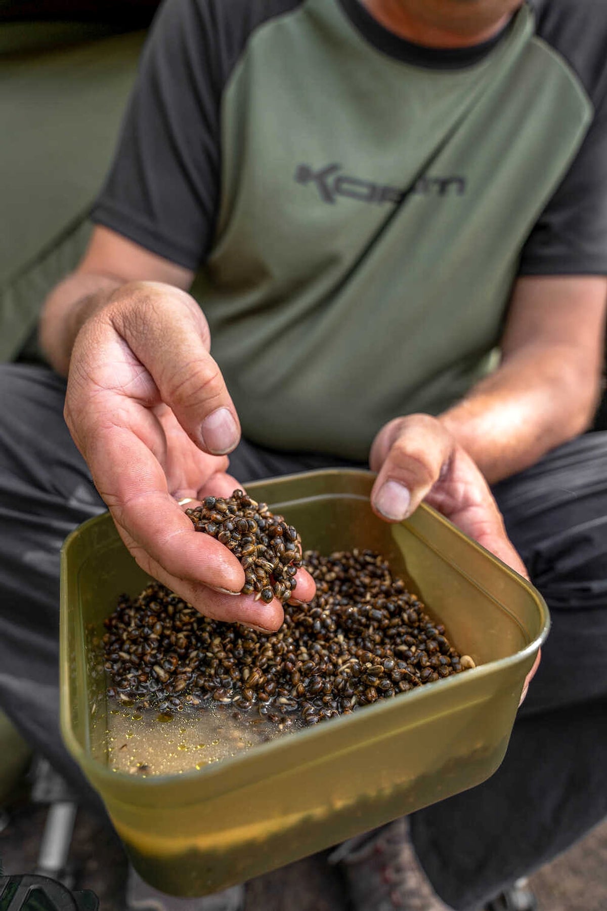 Visser schept hennepzaad uit een open Korum Bait Tub met olijfbruine wanden