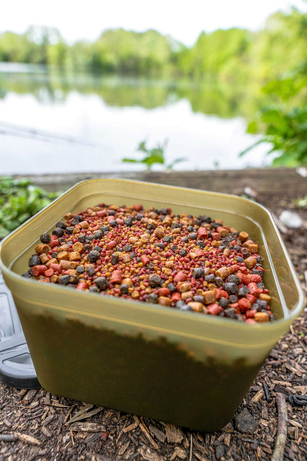 Open Korum Bait Tub gevuld met verschillende pellets op een boomstam naast het viswater
