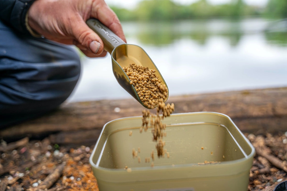 Korum voerschep laat bevochtigde pellets in de open Method Mix aasbak vallen aan de waterkant