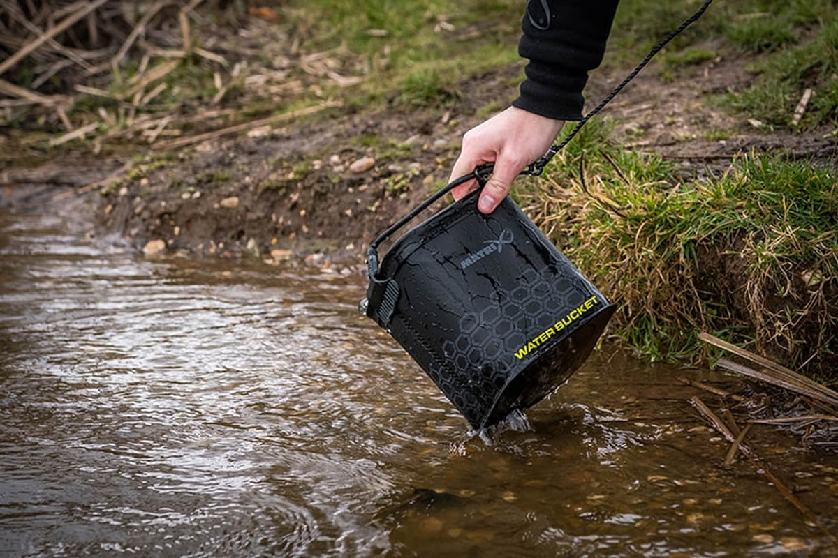 Een hand tilt de Matrix EVA Water Bucket aan de draaggreep op bij de oever