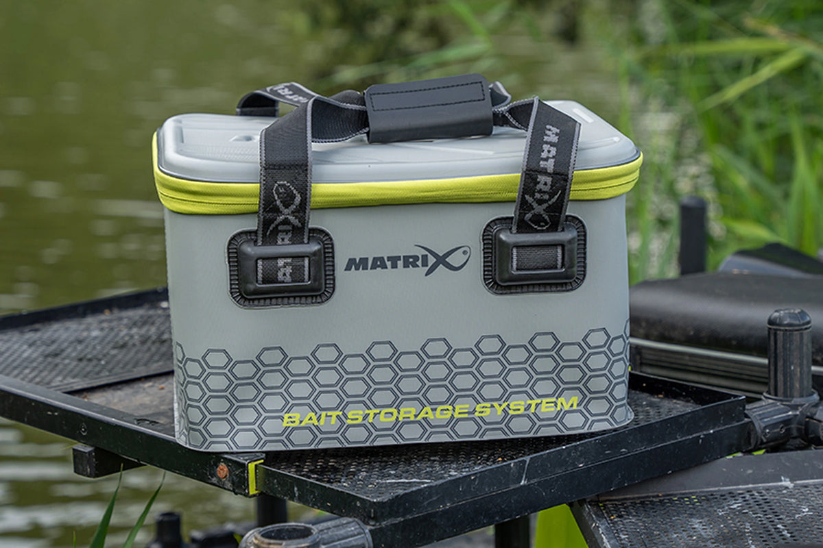 Matrix EVA Bait Storage System – de gesloten grijze aastas met gele rits op een zwarte zijtafel aan het water