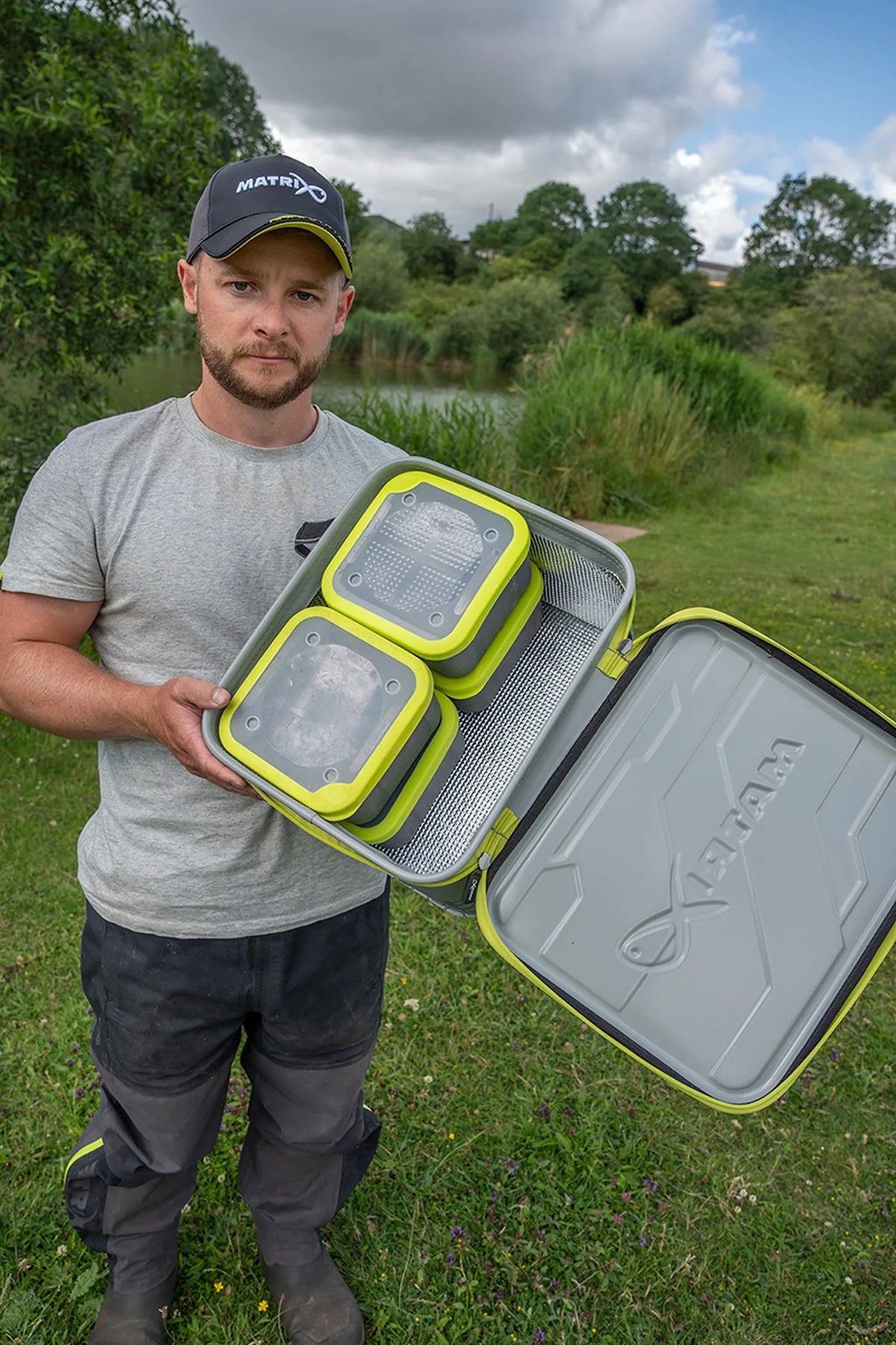 Matrix EVA Bait Storage System – visser toont de open aastas met grijze aasbakken met gele randen aan de waterkant
