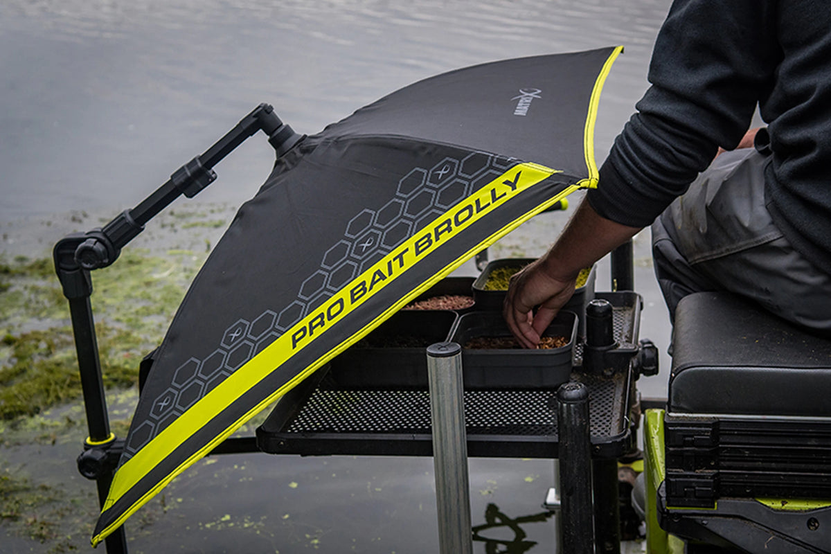 Matrix Pro Bait Brolly aasparaplu – visser neemt aas uit bakken onder de schuin geplaatste kap op de aastafel