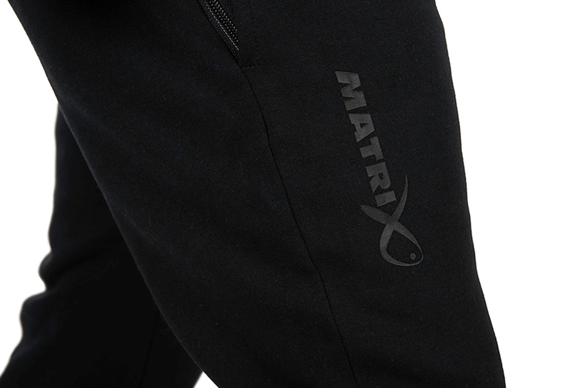 Matrix Sherpa Joggers – detail van het donkergrijze Matrix-logo en de zijnaad op de zwarte broekspijp