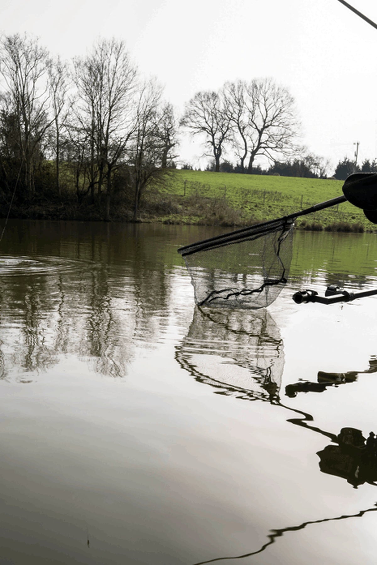 Preston Carbon Free Flow Landing Net boven het water, met het diepe gaasnet en de weerspiegeling in de vijver zichtbaar