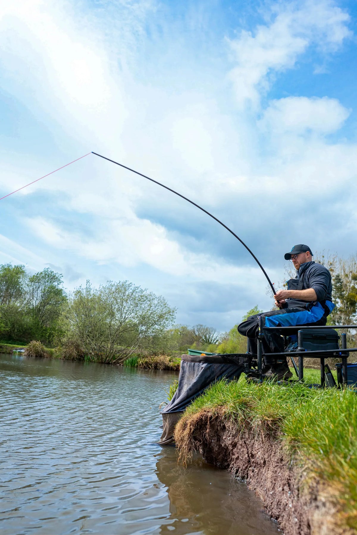 Visser aan de waterkant met een gebogen Preston DWA Kits Carp-topset en rood elastiek dat uit de hengeltop loopt