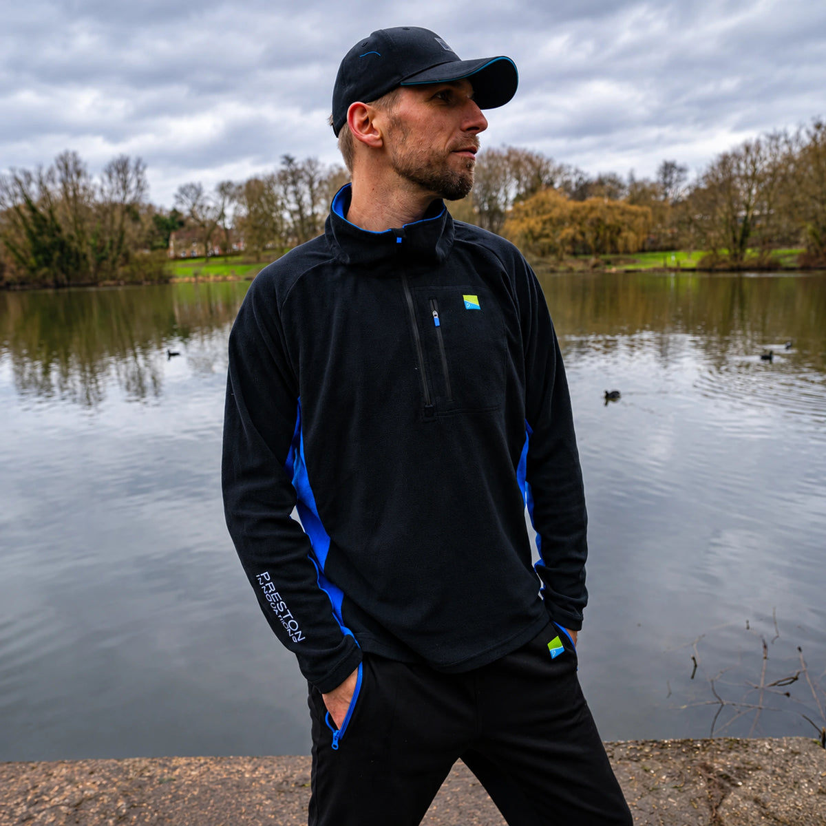 Preston Micro Fleece, gedragen aan de vijver in schuin vooraanzicht met blauwe zijpanelen en borstzak