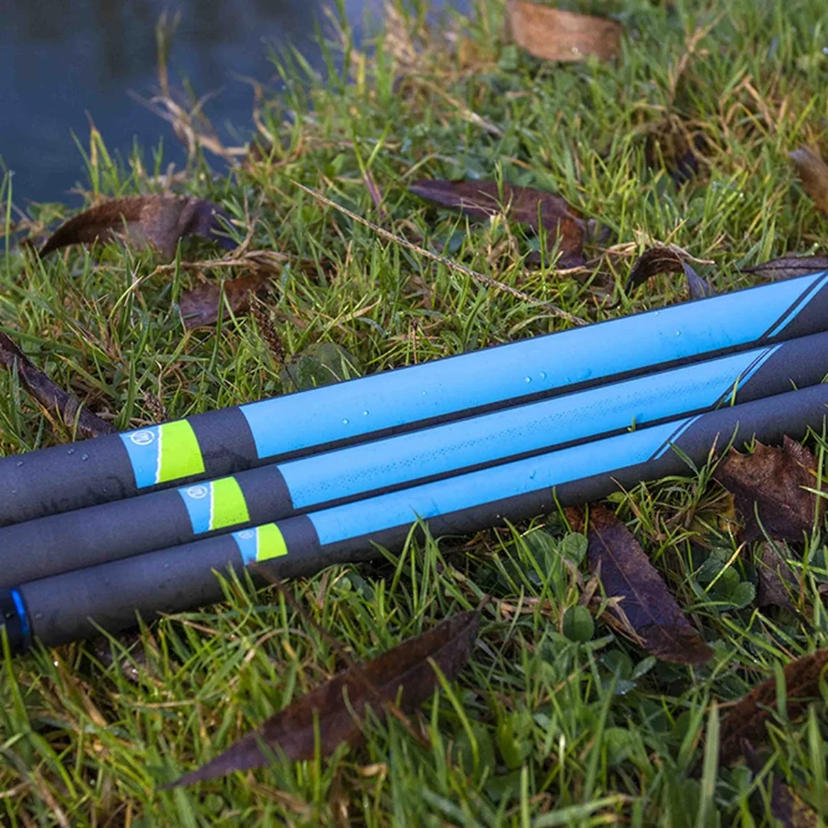 Drie Preston Response Whips met blauwe panelen op de handdelen, waterdruppels en groen-blauwe labels in het gras aan de waterkant