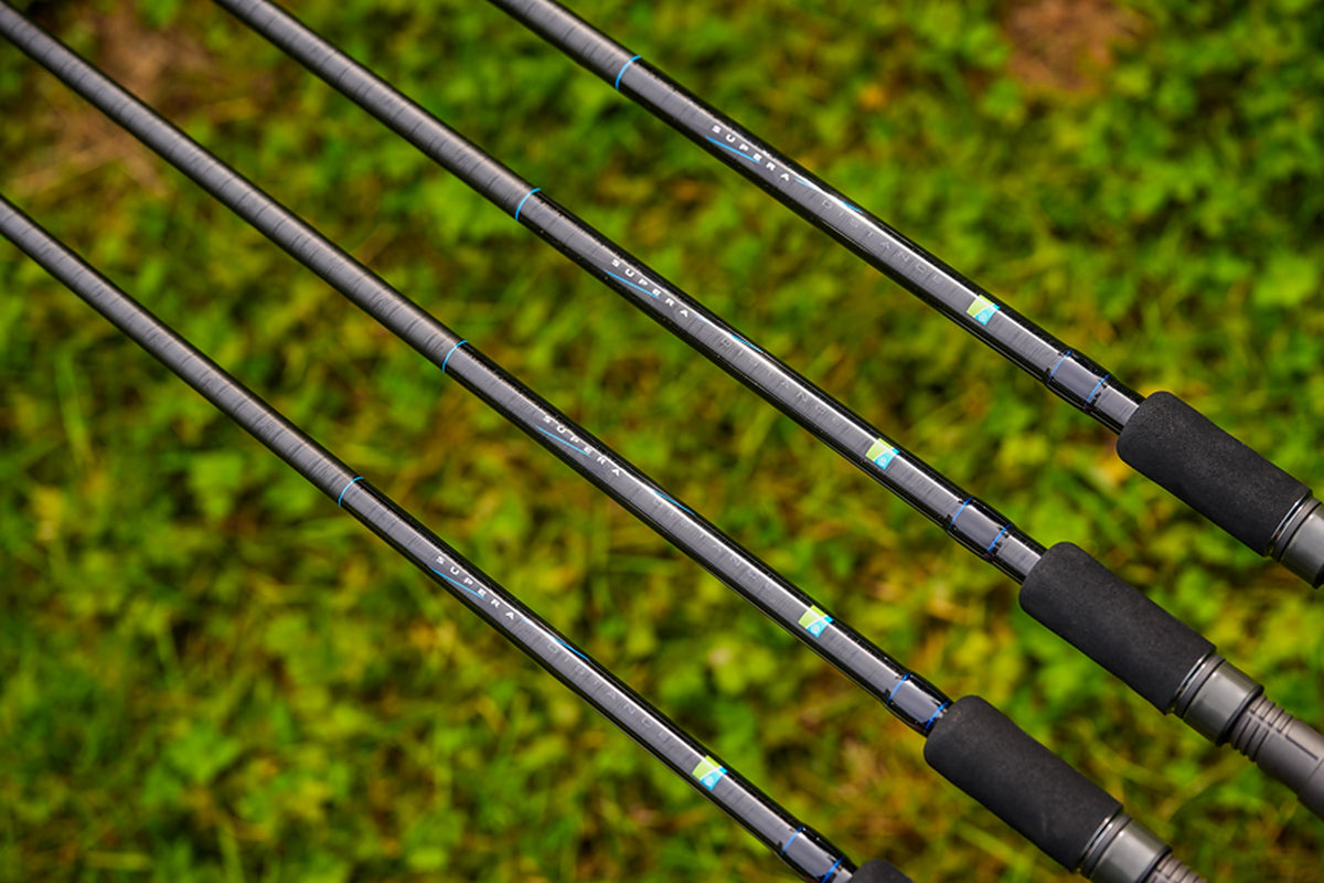 Preston Supera X Distance Feeder – vier hengelblanks met Supera-opdruk, blauwe sierwikkelingen en zwarte EVA-voorhandgrepen boven groen gras