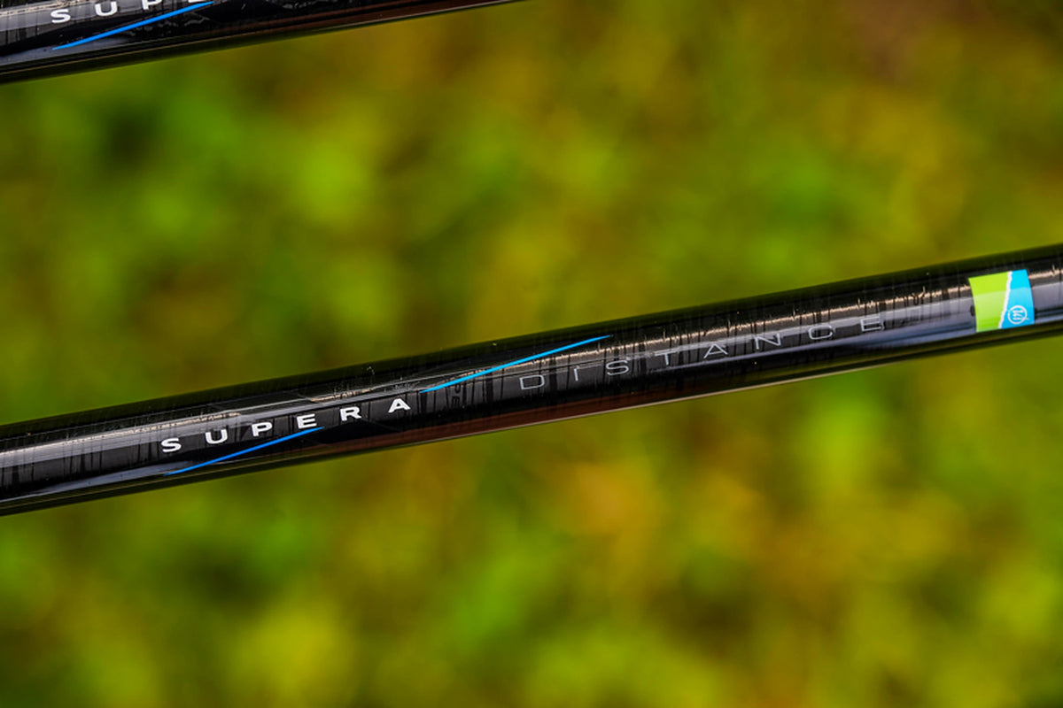 Preston Supera X Distance Feeder – detail van de SUPERA DISTANCE-opdruk en het groen-blauwe Preston-logo op de carbonblank tegen een groene achtergrond