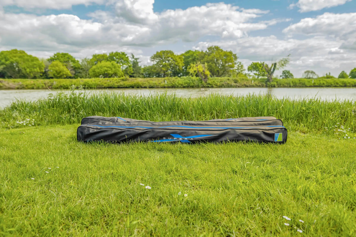 Preston Supera X Pole-foedraal liggend in het gras aan de waterkant, met lange blauwe rits en SUPERA-opdruk