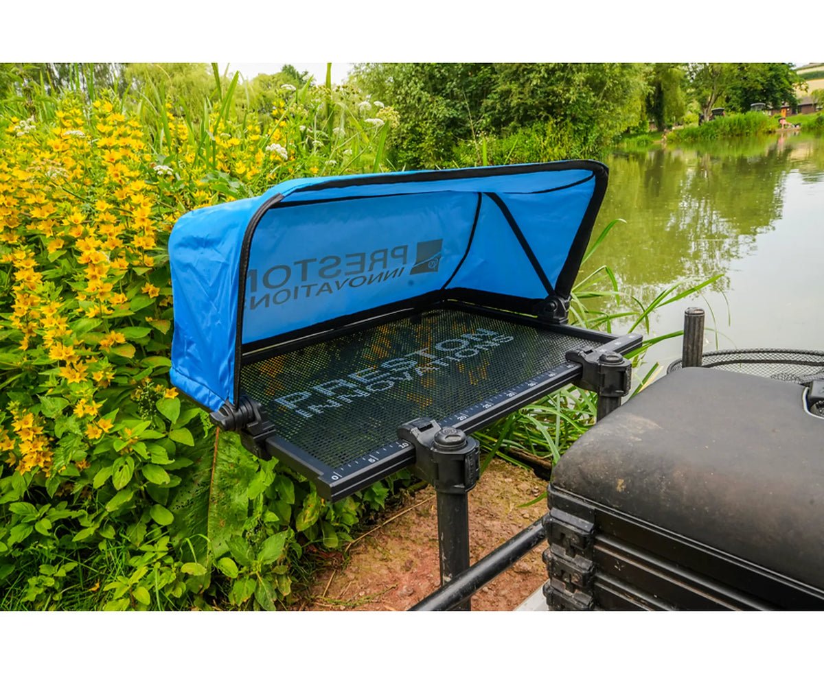 Preston Ventalite Side Tray XL – aastafel met blauwe beschermkap naast een zitkist aan het water