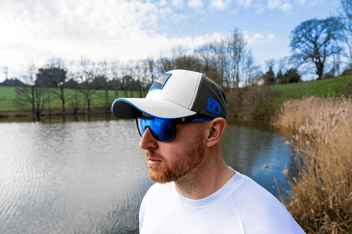 Preston White Drift Cap gedragen schuin van voren, met zonnebril en viswater op de achtergrond