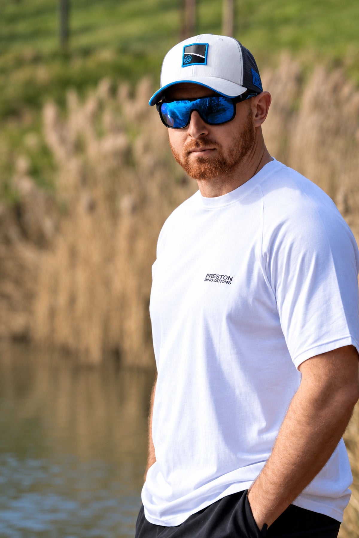 Preston White Drift Cap gedragen met zonnebril, in een staand portret bij de begroeide waterkant